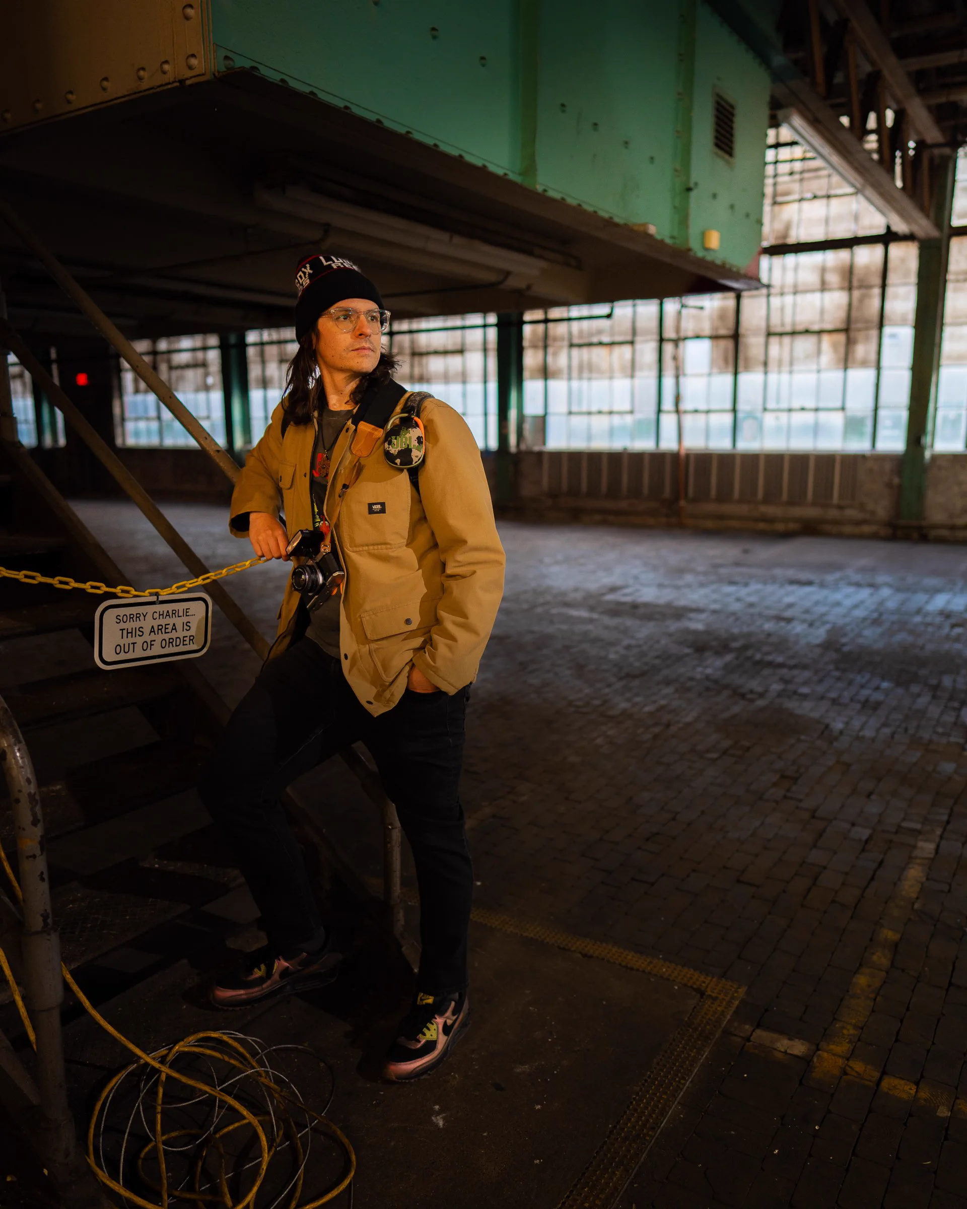 A photographer poses beside metal stairs and an out-of-order sign in an industrial hall.