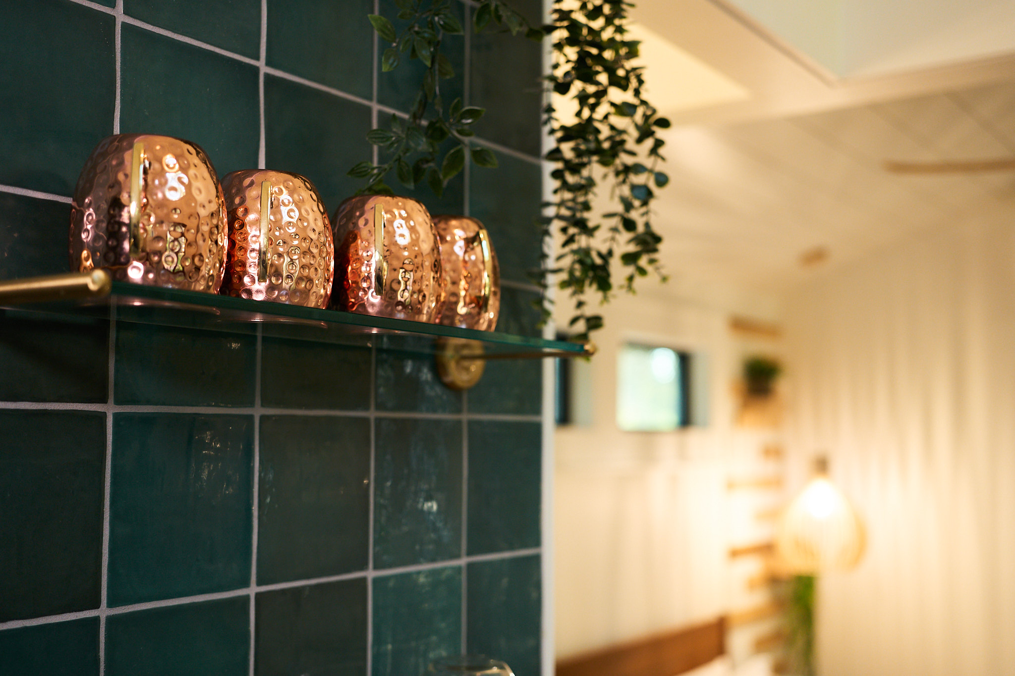 Copper mugs rest on a glass shelf against green tile.
