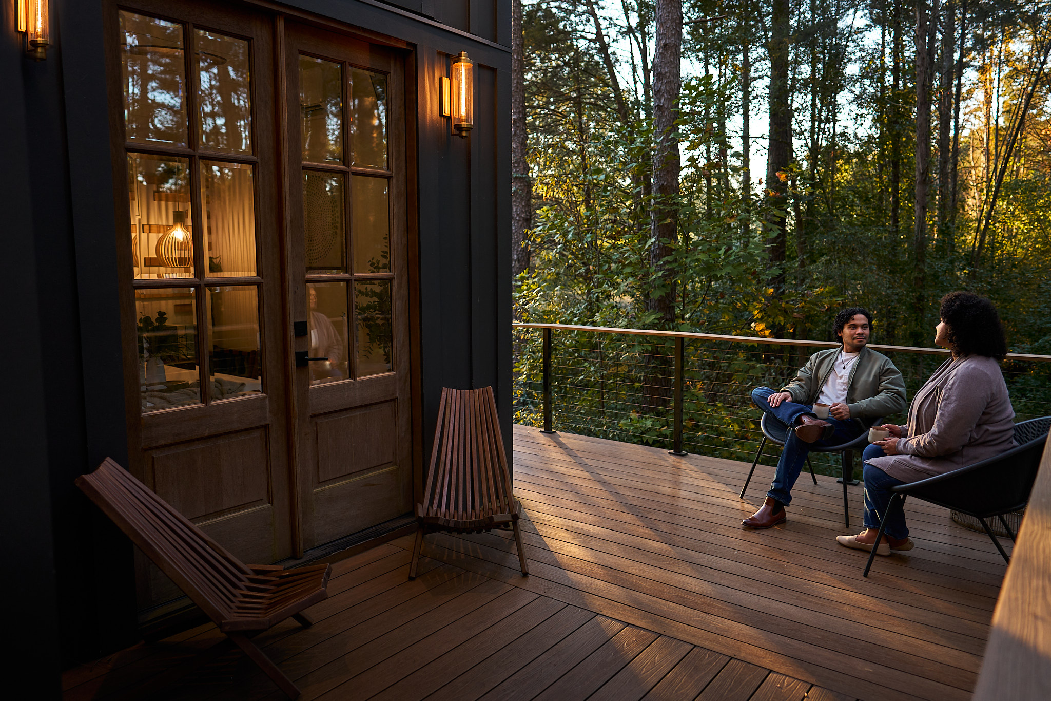 Two guests sit and talk on a treehouse porch surrounded by woods.