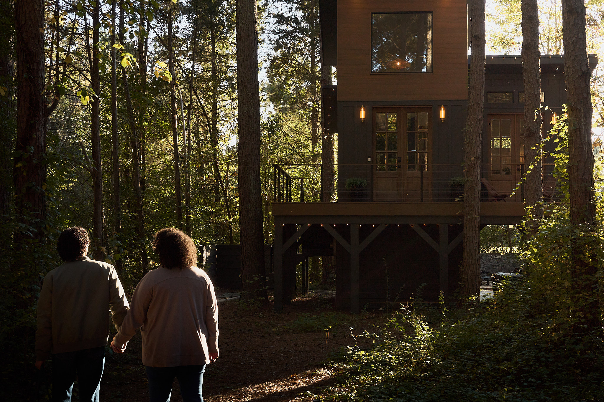A couple walks through the woods toward an elevated treehouse in warm afternoon light.